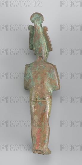 Figurine of Osiris depicted as a mummy-like standing figure wrapped in a shroud. The arms protruding from beneath the shroud are crossed over the chest in the so-called Upper Egyptian configuration. In his right hand, Osiris holds a scepter, while in his left he holds a whip. The head is adorned with an atef crown consisting of the White Crown and two ostrich feathers placed on the sides. The crown is topped with a solar disk and additionally adorned with a large uraeus with two lateral body coils and a long, wavy tail. The shroud fits softly to the body, highlighting the outline of the back and forearms. A false beard is attached to the chin. 664–332 BC; Late Period, Egypt; sculpture; height 17.9 cm, width 5 cm., A small metal statuette photographed against a pale gray background showing the back of a standing figure with a tall headdress; the surface is mottled with green and brown patina and areas of darker and lighter green, brown, and occasional reddish-brown; the figure has a nar...