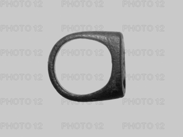 Ring, Ring with a band and a stone, cast from a single piece of metal. The band is narrow, convex on the outside and flat on the inside. The stone is arranged parallel to the line of the band. It is a long rectangle with rounded oval-shaped short edges. The flat surface of the stone is decorated with two hieroglyphic characters. 332–30 BC; Ptolemaic Period, Egypt. Jewelry; height 2.1 cm, diameter 2.3 cm., A single dark metal ring with an irregular, slightly squared profile sits centered against a light gray background; the ring's surface appears worn and textured with subtle highlights and shadows showing its rounded inner edge and thicker outer band, and the visible colors are shades of dark gray and light gray.