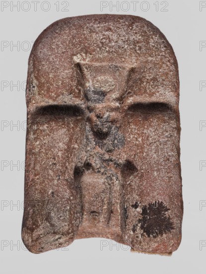 Mold for a figurine of the goddess Isis, Isis myth., Egyptian deities, Egyptian religion, crowns, 664–332 BC; Late Period, Egypt; mold; height 2.5 cm, width 5.1 cm, depth 3.4 cm, A weathered carved stone fragment with a rounded top and an irregular lower edge, showing a recessed central figure with a raised head and suggestions of shoulders and a torso, flanked by shallow grooves that create a cross-like negative space; the surface is rough and pitted with areas of flaking and granular texture, and a dark irregular blotch is visible near the lower right area. Colors visible include reddish-brown, beige, off-white, dark gray, and black.