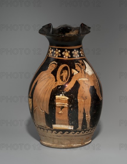 Red-figure oinochoe depicting a woman with a mirror and Eros, Apulia, late 4th century BC, vessel oinochoe, height 26.5 cm, width 17.5 cm, A ceramic vessel with a flared rim and rounded body sits against a neutral gray background; the vessel shows a palette of black, terracotta, beige, white, and hints of gold, with a band of floral motifs in light colors near the neck, a decorative wave pattern in dark and light near the base, and a painted scene on the bulbous body depicting a group of human figures in profile and three-quarter view, some draped in flowing garments, one holding a circular object, and a central rectangular pedestal-like form, with areas of worn and chipped surface revealing lighter underlying clay.