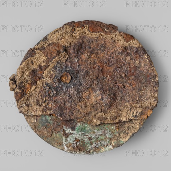 Cardiophilax – breastplate, Convex bronze disc decorated with an engraving of three concentric circles made by punching sheet metal on the reverse side. Engraving depicting a fantastic bird with two heads and four long legs. 2nd half of the 6th century BC. From Samnium, Southern Italy. Weapon/armor breastplate. Height 18 cm, width 20.5 cm, depth 2 cm, weight 0.344 kg., A circular, flat object sits against a light gray background, showing a rough, uneven surface with heavy corrosion and flaking. The visible area displays a mix of dark brown and reddish-brown rust textures, with patches of tan and beige crusty material layered across the top. Near the lower edge there are areas of green and pale green patina mixed with some white encrustation, and small spots of darker gray and black mottling scattered across the surface. The edges are irregular and chipped, and the surface has pitted, bubbly formations and a few raised, rounded nodules.