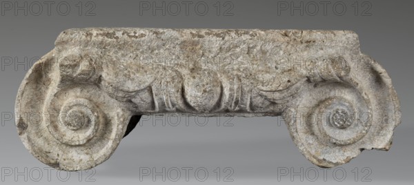 Ionic capital, unknown author, Roman architecture, capitals architect, volutes, ovoid ornament, cymatium ornament, Ionic capital with ovoid between volutes, 2nd–3rd century AD previously dated 1st half of 2nd – 1st half of 3rd century AD, Novae, Roman Empire 27 BC–476 — architectural element, element of architectural decoration, capital, architectural detail; height 18.5 cm, width 47.5 cm, depth 39 cm, weight 44 kg., A weathered stone fragment shaped like a horizontal architectural element with two symmetrical spiral scrolls at each end and a carved central band with rounded, leaf-like forms; the surface shows rough texture, chips, and cracks. The visible colors are shades of gray, off-white, beige, and light brown.