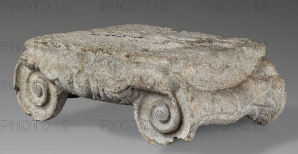 Ionic capital, Roman architecture, capitals architect, volutes, ovoid ornament, cymatium ornament, Ionic capital with ovoid between volutes, 2nd–3rd century AD previously dated 1st half of 2nd–1st half of 3rd century AD, found at Novae, Roman Empire 27 BC–476 — architectural element / element of architectural decoration, capital, architectural detail; height 18.5 cm, width 47.5 cm, depth 39 cm, weight 44 kg., The image shows a weathered stone fragment with a rough, textured surface in shades of gray, off-white, beige, and light brown, featuring a carved scroll motif at each visible corner and worn layered edges; the background is a smooth gradient of gray.