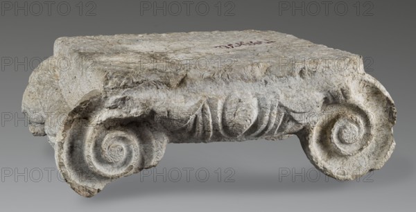 Ionic capital, Roman architecture, capitals architect, volutes, ovoid ornament, cymatium ornament, Ionic capital with ovoid between volutes, 2nd–3rd century AD formerly 1st half of 2nd – 1st half of 3rd century AD, Novae, Roman Empire 27 BC–476, architectural element / element of architectural decoration / capital / architectural detail; height 18.5 cm, width 47.5 cm, depth 39 cm, weight 44 kg, A weathered stone fragment shaped like a broken architectural element with a flat top surface and carved scroll-like volutes at each end rests against a neutral background; the stone shows rough, pitted texture and carved fluting along its front face, with visible chisel marks and irregular edges. The visible colors are shades of gray, off-white, beige, and subtle brownish tones, with slightly darker gray shadows around recessed areas. The background is a smooth gradient of gray.