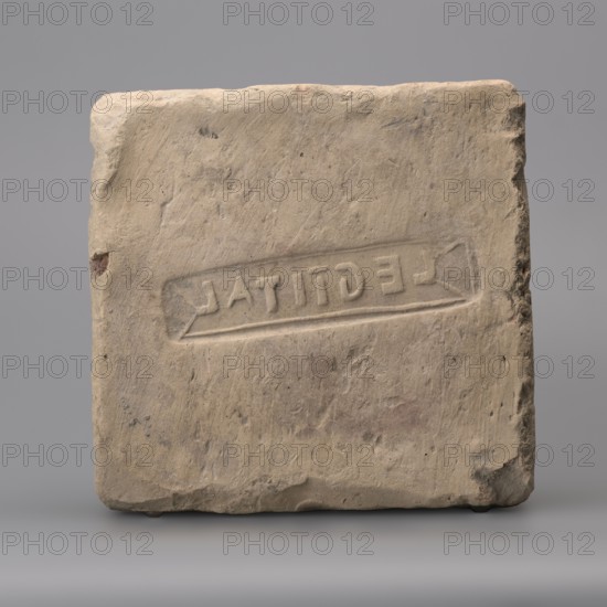 Hypocaust brick with stamp impression, square. From a hypocaust post. Negative stamp, 2nd half of the 3rd century, Novae; varia. Height 19 cm, width 19.5 cm, depth 8.5 cm, weight 4.16 kg., A roughly square stone block with a worn, textured surface sits against a neutral background; its color palette includes beige, tan, light brown, and hints of gray with small darker spots and patches, and the stone shows rough, chipped edges and indentations, with a shallow rectangular impression carved into the front face and subtle variations in shadow and surface roughness across the faces.
