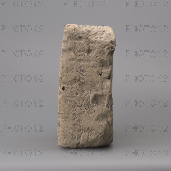 Hypocaust brick with stamp impression, square. From a hypocaust post. Negative stamp, 2nd half of the 3rd century, Novae, varia. Dimensions: height 19 cm, width 19.5 cm, depth 8.5 cm, weight 4.16 kg., A roughly rectangular, vertically oriented stone block with an uneven, weathered surface in shades of beige, tan, and light brown stands against a smooth gray background; the block shows pitted and rough textures, rounded and chipped edges, and several small holes and indentations, with subtle darker and lighter patches across its face creating a mottled appearance.