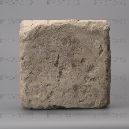 Hypocaust brick with stamp impression, square. From a hypocaust post. Negative stamp, 2nd half of the 3rd century, Novae, varia; height 19 cm, width 19.5 cm, depth 8.5 cm, weight 4.16 kg., A roughly square block of rough stone with an uneven, pitted surface, showing a weathered texture and small chips along its edges; the stone displays a range of earthy colors including beige, tan, light brown, gray, and hints of pale reddish tones, set against a smooth neutral gray background.