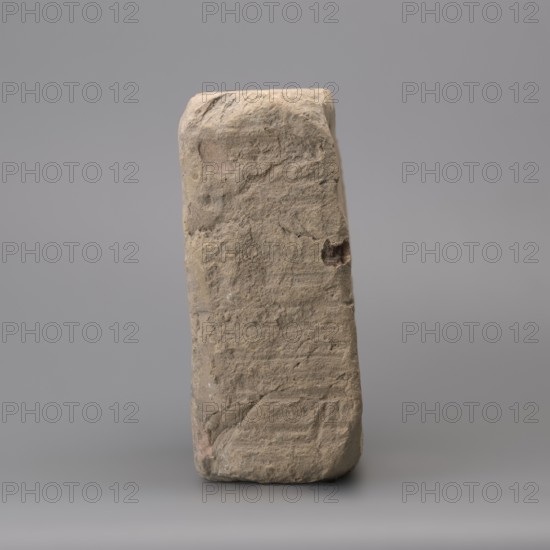 Hypocaust brick with stamp impression, square. From a hypocaust post. Negative stamp, 2nd half of the 3rd century, Novae; varia. Dimensions: height 19 cm, width 19.5 cm, depth 8.5 cm; weight 4.16 kg., A single upright rectangular stone block with rough, uneven surfaces and visible chips and indentations, shown against a neutral background; the stone displays a mix of beige, tan, light brown, and pale gray tones with subtle darker brown patches and slight pinkish undertones in some areas, and the background appears uniform gray.