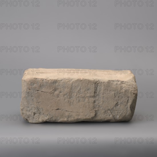 Hypocaust brick with stamp impression, square. From a hypocaust post. Negative stamp, 2nd half of the 3rd century, Novae; varia. Height 19 cm, width 19.5 cm, depth 8.5 cm, weight 4.16 kg., A roughly rectangular, worn brick or stone block sits on a smooth surface against a plain background; the object shows a textured, uneven surface with chips, dents, and rough patches, and its colors include shades of beige, tan, light brown, and some grayish tones, while the surrounding surface and background are various shades of gray.