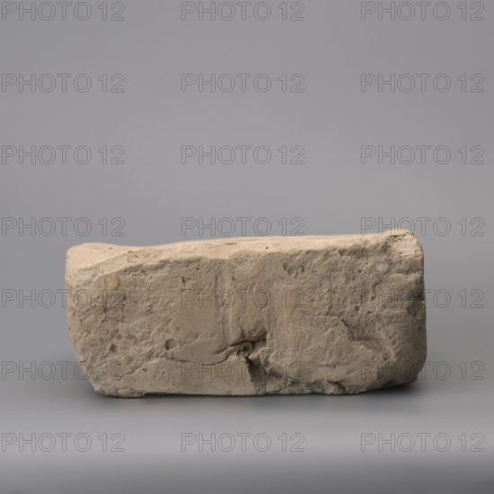 Hypocaust brick with stamp impression, square. From a hypocaust post. Negative stamp, 2nd half of the 3rd century, Novae, varia. Height 19 cm, width 19.5 cm, depth 8.5 cm, weight 4.16 kg, A rectangular rough stone slab with an uneven, weathered surface sits against a smooth backdrop; the slab shows a variety of textures including pitted areas, small cracks, and a larger chipped section near its base, with subtle rounded edges and a slightly sloping top. The visible colors are beige and light tan on the stone, with hints of darker brown in crevices, and the background and surface beneath the slab are shades of gray.