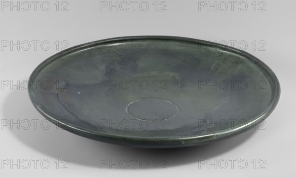 Plate, shallow stone plate with a convex bottom and a strongly flaring bowl; wide rim bent inward. Ca. 3100–2686 BC, Early Dynastic Period; found at Edfu. Vessel, height 5.6 cm, diameter 40 cm., A shallow, round dish with a gently raised rim is shown, displaying a smooth surface with visible hairline cracks and repaired seams; the dish is primarily dark green with areas of lighter green and subtle bluish-green tones, and there are a few darker gray-green patches and faint highlights that catch the light.