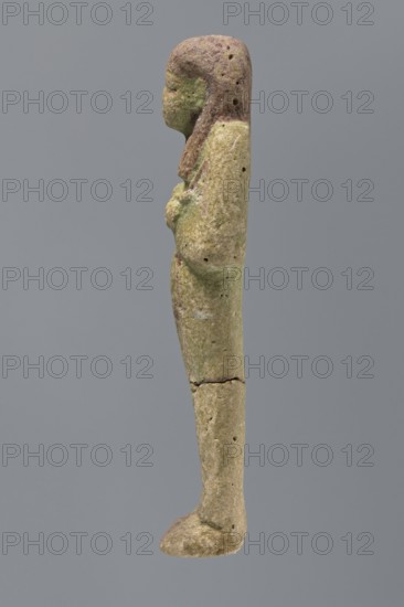 Ushabti ?-en-Khonsu, Mummy-shaped figurine shabti, funerary gift, hoes; arms crossed on the chest, right folded over left, holding hoes in both hands. Convex tools; rectangular basket low on the back. Smooth tripartite wig; large ears; facial details poorly legible. Wig, facial details and basket painted black. Painted inscription in a framed column on the front of the legs: Osiris the god father of Amun, Waad -en-Khonsu, scribe, justified by the voice. c. 1069–c. 945 BCE; Third Intermediate Period, 21st Dynasty, Egypt. Height 12.7 cm., A slender vertical figurine viewed in profile against a solid gray background, with a rounded head and simplified facial outline, a long neck and a straight torso leading into an elongated leg and foot; the surface shows a rough, pitted texture with small holes and a visible horizontal crack at the knee area; colors visible include muted green, pale beige, brown, and hints of darker brown on the head and upper body.