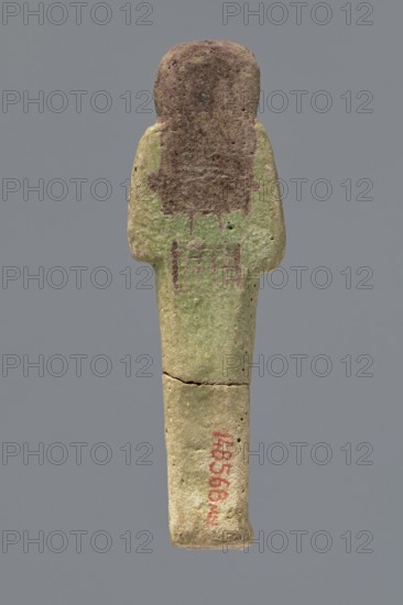 Ushabti ?-en-Khonsu, Mummy-shaped figurine shabti. Arms crossed on the chest, right folded over left, holding hoes in both hands; convex tools. Rectangular basket low on the back. Smooth tripartite wig, large ears; facial details poorly legible. Wig, facial details and basket painted black. Painted inscription in a framed column on the front of the legs: Osiris the god father of Amun, Waad-en-Khonsu, scribe, justified by the voice. c. 1069–c. 945, Third Intermediate Period, 21st Dynasty, Egypt. Height 12.7 cm., A single upright carved figure with a rounded head and elongated body, showing a worn surface texture and small holes, colored in shades of green and brown with areas of darker brown on the head and upper torso and traces of red on the lower body; the background is a solid gray.