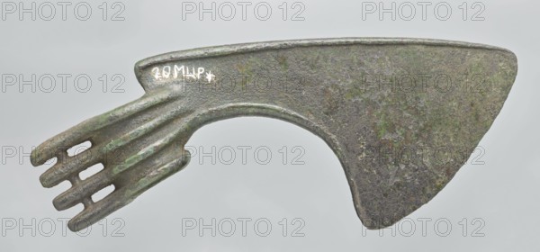 Battle axe, unknown Luristan workshop. Battle axe with a low cylindrical socket with a round hole; a blade grows from the socket terminating in a semicircular cutting edge set at ~45° to the shaft axis. The base of the blade protrudes above the socket; its upper edge is sloping and straight with a slight central bulge, the lower edge is shorter and a slightly obtuse arc. Four identical blunt-tipped spikes protrude from the butt, connected vertically at mid-length; the spikes extend into side ribs on both sides of the socket — the upper two terminate at the base of the blade, the third extends into its lower edge, and the fourth runs around the lower edge of the socket reinforcing it. Bronze Luristan weapon, 2nd half of the 2nd millennium BC; Iran Luristan. Weight 334 g; height 10.2 cm; length 19 cm; thickness 3.2 cm., An aged metallic object with a textured surface showing a patina of green and brown tones, featuring a broad triangular plate on one end and a curved arch connecting to a...