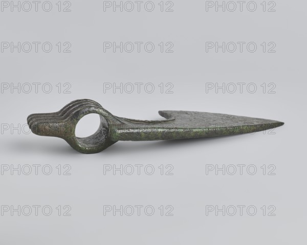 Battle axe, unknown Luristan workshop. Battle axe with a low cylindrical socket with a round hole, from which grows a blade terminating in a semicircular cutting edge set at ~45° to the shaft axis. The base of the blade protrudes above the socket; its upper edge is sloping and straight with a slight central bulge, the lower edge is shorter and a slightly obtuse arc. Four identical blunt-tipped spikes protrude from the butt, connected vertically at mid-length. The spikes extend into side ribs on both sides of the socket: the upper two terminate at the base of the blade, the third extends into its lower edge, and the fourth runs around the lower edge of the socket, further reinforcing it. Luristan bronzes; weapon. 2nd half of the 2nd millennium BC. Iran; Luristan. Weight 334 g; height 10.2 cm; length 19 cm; thickness 3.2 cm., A single metallic object lies on a pale gray background, showing a long flat blade that tapers to a pointed tip and a rounded section with a circular hole near the ...