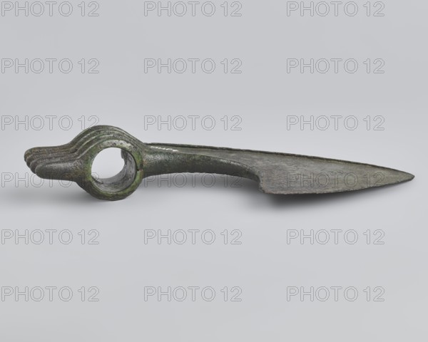 Battle axe, unknown Luristan workshop. Battle axe with a low cylindrical socket with a round hole, from which grows a blade terminating in a semicircular cutting edge set at about 45° to the shaft axis. The base of the blade protrudes above the socket; its upper edge is sloping and straight with a slight central bulge, the lower edge is shorter and slightly obtuse-arched. Four identical blunt-tipped spikes protrude from the butt, connected vertically at mid-length; the spikes extend into side ribs on both sides of the socket — the upper two terminate at the base of the blade, the third extends into its lower edge, and the fourth runs around the lower edge of the socket for reinforcement. Luristan, Iran; 2nd half of the 2nd millennium BC. Bronze weapon. Weight 334 g; height 10.2 cm; length 19 cm; thickness 3.2 cm., A metal object with a long flat blade-like section tapering to a pointed end lies horizontally on a pale gray background; the opposite end forms a rounded ring with a hollow ...