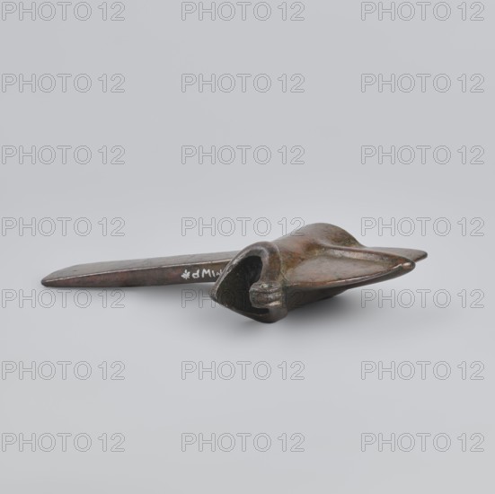 Battle axe, Eastern Gallic Scenography, 2nd half of 3rd millennium BC, Western Iran, weapon, weight 280 g, A bronze object with a flat elongated handle and a wider, curved end resembling a stylized head or scoop lies on a neutral light gray background; the object shows a patina with brown, green, and darker bronze tones and subtle highlights, and a few areas of shinier metallic reflection are visible on its surfaces.