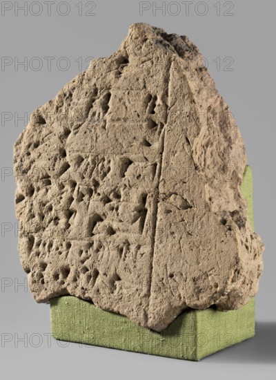 Brick fragment with Gudea's foundation inscription, Gudea ca. 2150 BC, architectural element with cuneiform writing, Near Eastern architecture, ca. 2150 BC 2nd Dynasty of Lagash, found at Tello, origin Girsu Sumer, Tello, Iraq, foundation inscription, weight 1.35 kg, A roughly triangular, textured stone slab with an eroded, pitted surface rests on a rectangular fabric-covered support; the stone is primarily tan and beige with subtle darker brown shadowing in crevices, and the support is a muted green. The slab shows a variety of small cavities and raised ridges across its face, with an uneven, jagged top edge and a more vertical, straighter right edge; the lower edge sits flush against the green fabric base. The background is a smooth, neutral gray that contrasts with the granular texture of the stone and the woven texture of the green support.