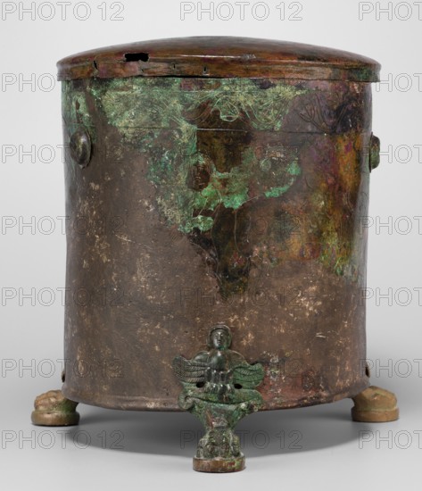 Cista container decorated with a depiction of the Dioscuri and Athena. Cylindrical sheet-metal vessel on three legs, engraved with the Dioscuri and Athena, 8 figures preserved. Bands of engraved plant decoration under the rim and at the lower edge. Concave bottom. Legs shaped like predator paws ending in sirens. Slightly convex sheet-metal lid engraved with two hippocampi and a trace of a central handle. Dated 4th century BC, possibly Praeneste Italy. Vessel; weight 1.132 kg; height 24.5 cm; bottom diameter 20 cm; top diameter 22 cm., A cylindrical metal container with a domed lid stands on a plain surface, showing aged and worn surfaces with patches of green and turquoise patina, areas of dark brown and reddish-brown metal, and some faint purple and ochre discoloration. The lid has an irregular dark opening and a slightly raised rim with darker brown and reddish tones. The body of the container displays mottled green corrosion and darker brown areas, with decorative circular protrusio...