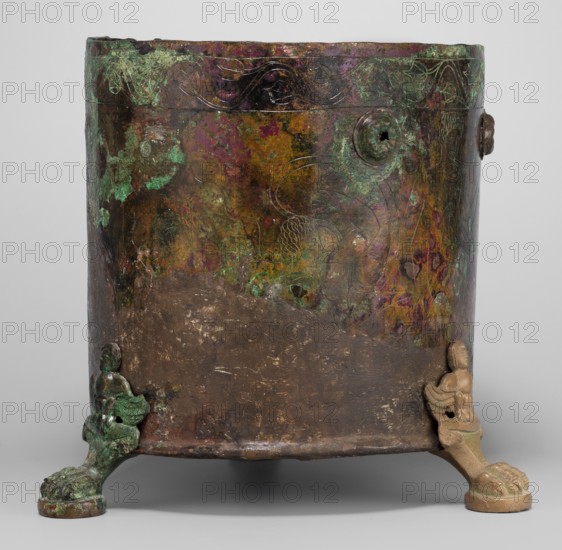 Cista container decorated with a depiction of the Dioscuri and Athena. Cylindrical vessel made of sheet metal, decorated with an engraved depiction of the Dioscuri and Athena, 8 figures preserved. Under the rim and at the lower edge are bands of engraved plant decoration. Concave bottom. Legs in the shape of predator's paws, ending in sirens. Slightly convex lid, made of sheet metal, decorated with an engraved depiction of two hippocampi. Trace of a handle in the centre. Dates: 4th century BC; 2nd half of the 18th century later intervention. Place of origin: Praeneste Italy. Type: metal vessel, cista on 3 legs. Weight 1.132 kg; height 24.5 cm; diameter 20 cm bottom, 22 cm top., A cylindrical metal vessel standing on a pair of decorative tripod legs, with a weathered surface showing a mottled patina in green, brown, purple, and hints of gold; the upper rim has a band with faint engraved curving motifs, while the body displays areas of darker brown wear and irregular splotches of green a...