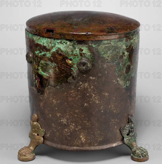 Cista container decorated with a depiction of the Dioscuri and Athena. Cylindrical vessel made of sheet metal, decorated with an engraved depiction of the Dioscuri and Athena, 8 figures preserved. Under the rim and at the lower edge are bands of engraved plant decoration. Concave bottom. Legs in the shape of predator's paws, ending in sirens. Slightly convex lid, made of sheet metal, decorated with an engraved depiction of two hippocampi. Trace of a handle in the centre. 4th century BC, Praeneste Italy. Vessel; metal; weight 1.132 kg; height 24.5 cm; diameter 20 cm bottom, diameter 22 cm top., A cylindrical metal container with a rounded lid stands on a neutral background, showing a mottled surface of dark brown and green patina with areas of bright green corrosion and touches of reddish-brown; the lid is darker with subtle variations and a small hole at the rim. The body features a few raised circular rivet-like elements and traces of etched or scratched linear patterns amid the green...