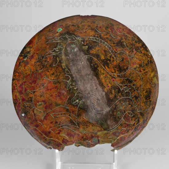 Cista container decorated with a depiction of the Dioscuri and Athena. Cylindrical vessel made of sheet metal, decorated with an engraved depiction of the Dioscuri and Athena, 8 figures preserved. Under the rim and at the lower edge are bands of engraved plant decoration. Concave bottom. Legs in the shape of predator's paws, ending in sirens. Slightly convex lid, made of sheet metal, decorated with an engraved depiction of two hippocampi. Trace of a handle in the centre. Cista on 3 legs. Metal vessel. Classified Gallic scenics; Athena mythology; Dioscuri mythology. 4th century BC, Praeneste Italy. Vessel; weight 1.132 kg; height 24.5 cm; diameter 20 cm bottom, 22 cm top., A round, weathered metal disc with a patina of green, brown, orange, red, and dark brown tones, showing a lighter vertical streak near the center; faint incised line decorations form flowing, curved motifs and stylized shapes across the surface; the disc edge is irregular with small chips and a small hole near the rim...