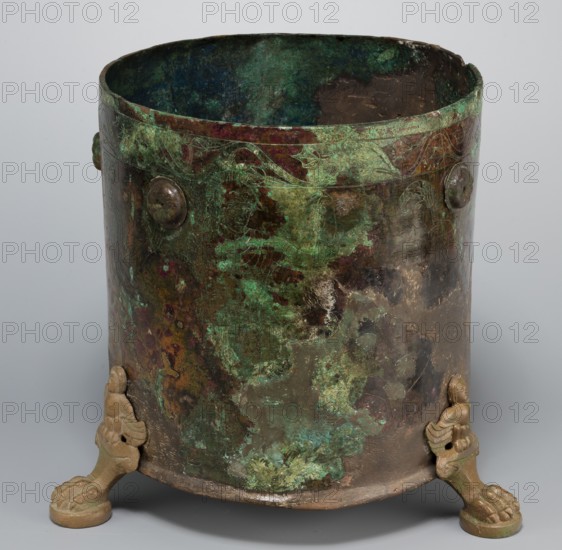 Cista container decorated with a depiction of the Dioscuri and Athena. Cylindrical vessel made of sheet metal, decorated with an engraved depiction of the Dioscuri and Athena, 8 figures preserved. Under the rim and at the lower edge are bands of engraved plant decoration. Concave bottom. Legs in the shape of predator's paws, ending in sirens. Slightly convex lid, made of sheet metal, decorated with an engraved depiction of two hippocampi. Trace of a handle in the centre. Dating: 4th century BC, Praeneste Italy. Metal vessel, cista on 3 legs. Weight 1.132 kg; height 24.5 cm; diameter 20 cm bottom, 22 cm top., A cylindrical metal vessel with a weathered surface showing a mix of green, dark green, brown, bronze, and hints of bluish tones, resting on a plain neutral background. The rim is irregular and the interior shows dark green and bluish patina. The exterior surface has mottled patches of green corrosion and darker brown areas, with a few raised circular rivet-like elements visible ne...