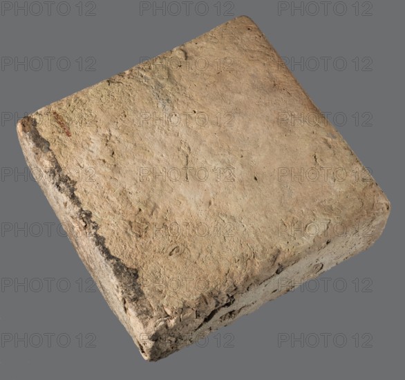 Brick with an inscription of King Shalmaneser III, Shalmaneser III 858–824 BC Shulmanu-ašared, king of the Neo-Assyrian Empire. From Calah Kalhu, the ziggurat at Kalhu commonly known as Nimrud. Square brick inscribed on the upper surface with seven lines of carefully impressed cuneiform characters. Sand-colored clay, one edge burnt to black. Date: 858–824 BC. Material: brick. Weight: 20.97 kg; width: 35 cm; length: 35 cm; thickness: 12 cm., A single rectangular block with worn, rough surfaces and rounded corners resting against a plain background; the block shows a textured, uneven top and sides with visible chips and small indentations, and a darker irregular strip along one edge. The colors visible are beige, tan, light brown, gray, and a few darker brown and black areas.