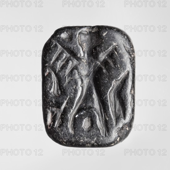 Rectangular stamp seal with a handle in the form of a protruding ridge along the axis of the seal with a crosswise drilled hole. On a rectangular base with rounded corners is a schematic representation of a demon, in human form and with wings instead of arms. Unspecified filler elements are on the sides of the demon. Stamp seal; c. 1650–c. 1350 BC Late Bronze Period. Made in Northwest Syria; found in the Near East. Height 1.7 cm, width 4.2 cm, depth 3.2 cm., A small rounded rectangular object with a dark glossy surface showing carved shallow grooves and raised ridges that form an abstract pattern; the object appears mostly black with a few small pale spots and lighter scuffed areas, and it sits against a plain light gray background.
