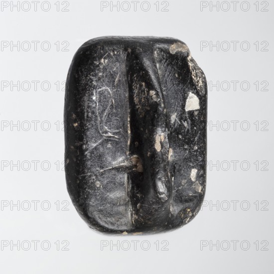 Rectangular stamp seal with a handle in the form of a protruding ridge along the axis of the seal with a crosswise drilled hole. On a rectangular base with rounded corners is a schematic representation of a demon, in human form and with wings instead of arms. Unspecified filler elements are on the sides of the demon. Materials/technique: seal. Date: c. 1650–c. 1350 BC Late Bronze Period. Place of manufacture: Northwest Syria; find region: Near East. Dimensions: height 1.7 cm, width 4.2 cm, depth 3.2 cm., A single smooth, irregularly shaped dark object with a mostly black surface and a few lighter spots and patches, showing worn and scratched areas, shallow grooves, and a central vertical depression. The object has a rounded rectangular outline with softer edges and a subtle glossy sheen, and the visible colors include black, white, and gray.