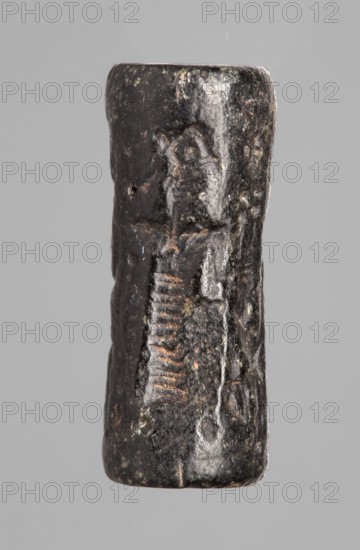 Cylinder seal with a mythological scene, unknown Cypriot workshop; mythological scene depicting two human figures dressed in long, crosswise pleated robes, standing opposite a winged lion standing on its back. The first figure holds the lion's lowered left front paw with its outstretched right hand; its left hand is lowered. The second figure, standing behind the first, holds a four-legged animal hanging upside down by its hind legs in its right hand; its left hand is lowered and extended towards the lion's hind legs. The animal is squeezed into a narrow space between the standing figures. Fantasy creatures, winged lions. 14th century BC; Late Bronze Period previously dated 15th–14th century BC. Near East; Cyprus. Seal. Height 2.55 cm; width 1 cm., A small cylindrical object with a rough, worn surface shown against a plain gray background; the object appears predominantly dark brown to black with areas of lighter brown and occasional grayish highlights from reflective wear; its surface...