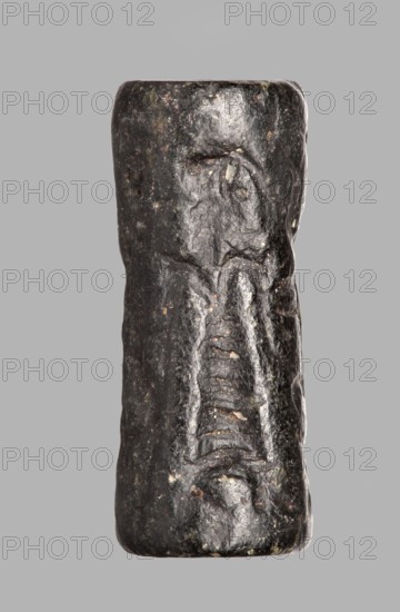 Cylinder seal with a mythological scene, unknown Cypriot workshop; mythological scene depicting two human figures dressed in long, crosswise pleated robes, standing opposite a winged lion standing on its back. The first figure holds the lowered left front paw of the lion with its outstretched right hand; its left hand is lowered. The second figure, standing behind the first, holds a four-legged animal hanging upside down by its hind legs in its right hand; its left hand is lowered and extended towards the lion's hind legs. The animal is squeezed into a narrow space between the standing figures. Fantasy creatures, winged lions. 14th century BC; Late Bronze Period previously dated 15th–14th century BC. Near East, Cyprus. Seal; height 2.55 cm, width 1 cm., A dark cylindrical stone object with a slightly irregular, worn surface set against a neutral gray background, showing a carved vertical motif resembling an abstract figure with rounded upper forms and a recessed central area with horiz...