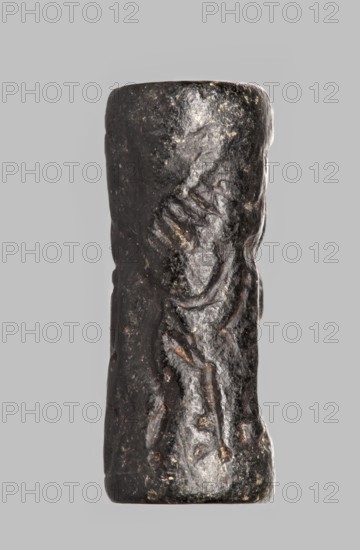 Cylinder seal with a mythological scene, unknown Cypriot workshop, fantasy creatures, mythological scene depicting two human figures dressed in long, crosswise pleated robes, standing opposite a winged lion standing on its back. The first figure holds the lowered left front paw of the lion with its outstretched right hand; its left hand is lowered. The second figure, standing behind the first, holds a four-legged animal hanging upside down by its hind legs in its right hand; its left hand is lowered and extended towards the lion's hind legs. The animal is squeezed into a narrow space between the standing figures. 14th century BC; Late Bronze Period previously dated 15th–14th century BC. Near East; Cyprus. Seal. Height 2.55 cm, width 1 cm., A vertical cylindrical object with a rough, uneven surface, dark brown to black in color with a few lighter brown and grayish patches, showing shallow carved or worn grooves and irregularities along its length, set against a plain light gray backgrou...