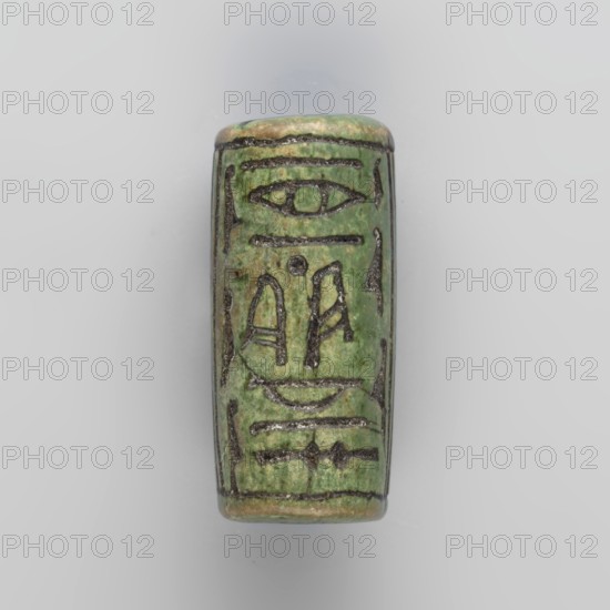 Cylinder seal with pseudo-hieroglyphs, A cylindrical seal with a hole drilled along the cylinder. Slightly more than half of the surface between two parallel border lines is occupied by a vertical band of hieroglyphic inscription delimited on the sides by a double line. 19th century, seal, height 2.6 cm, width 1 cm, A cylindrical bead-like object with a smooth, slightly glossy surface in shades of green and pale tan, decorated with several incised linear and curved markings filled with darker pigment, with edges showing worn tan and brown tones; the background is a neutral light gray.