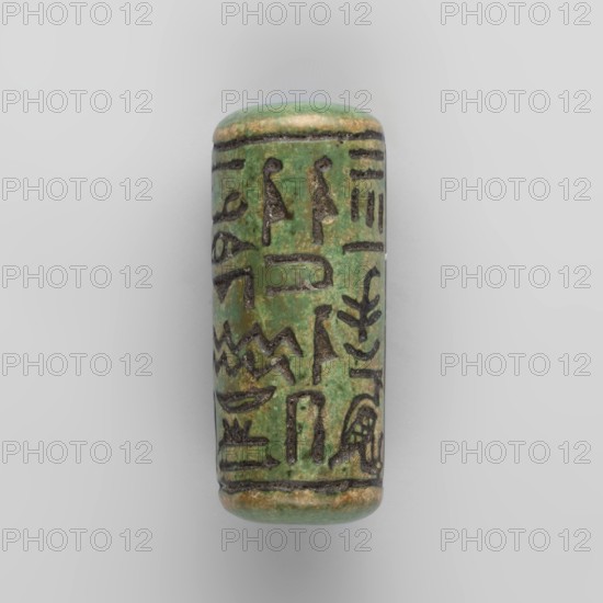 Cylinder seal with pseudo-hieroglyphs;, Cylindrical seal with a hole drilled along the cylinder. Slightly less than half of the surface between two parallel border lines is occupied by a vertical band of hieroglyphic inscription delimited on the sides by a double line. 19th century, origin unknown. Seal; height 2.5 cm, width 1.1 cm., A small cylindrical object with rounded ends sits against a light gray background; its surface is predominantly green with areas of brownish-beige and darker worn spots, and it is marked with several dark incised linear and pictorial motifs arranged vertically around the cylinder, creating a repeating decorative band.