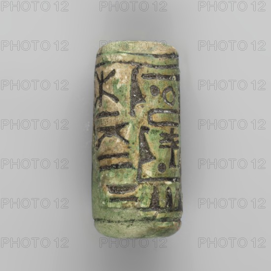 Cylinder seal with pseudo-hieroglyphs. A cylindrical seal with a hole drilled along the cylinder. The seal is covered with a greenish glaze along the vertical axis. On one side of the cylinder, an engraved hieroglyphic inscription is visible, enclosed in a frame. The hieroglyphic signs were filled with black dye. At the top and bottom, the cylinder is surrounded by an engraved ring. 19th century, unknown place of origin. Seal; height 2.7 cm, width 1.2 cm., A cylindrical object with a worn, uneven surface showing shades of green, beige, and dark brown, decorated with carved linear and geometric patterns filled with darker material, photographed against a light gray background.