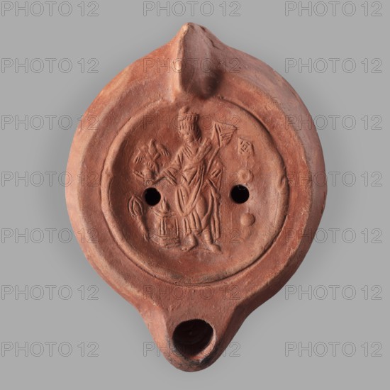 Lamp decorated with a representation of the goddess Roma with an eagle and a palm branch; at the goddess's foot a burning altar and a crab. 2nd century, Carthage Tunisia. Oil lamp, weight 0.152 kg. Single-burner lamp, Roman deities., A round terracotta object with a spout and a rounded pouring lip, shown against a gray background; the surface is reddish-brown with variations of darker brown and small worn areas, and there is a shallow circular recessed panel on top containing a low-relief depiction of a standing figure holding a staff and wearing draped clothing, with a smaller object beside the figure and a group of rounded holes puncturing the panel, plus a few small raised bumps and subtle tool marks on the textured surface.