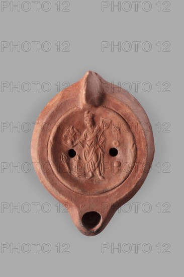 Lamp decorated with a representation of the goddess Roma with an eagle and a palm branch; at the goddess's foot a burning altar and a crab. Roman single-burner oil lamp, 2nd century, Carthage Tunisia. Weight 0.152 kg., A worn terracotta object with a rounded body and a pointed spout is shown against a gray background, displaying a recessed circular area with a raised central scene depicting a standing robed figure holding objects and a small raised motif nearby; the surface shows a few circular holes and a small hollow at the spout, with the clay surface exhibiting reddish-brown and muted orange tones along with darker brown patches and slight variations in texture and wear.