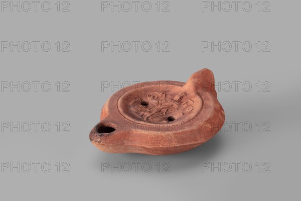 Lamp decorated with a representation of the goddess Roma: single-burner oil lamp depicting Roma with an eagle and a palm branch; at the goddess's foot a burning altar and a crab. Roman deities, 2nd century, Carthage Tunisia. Weight 0.152 kg., A small shallow clay oil lamp in a reddish-brown color rests on a smooth gray surface, with a rounded body and a raised rim surrounding a central recessed area that contains low-relief decorative motifs; the lamp has a short pointed nozzle with a darkened tip and an opposite small upright handle-like projection, and the clay surface shows variations of lighter and darker reddish-brown with some areas of darker sooting near the nozzle.