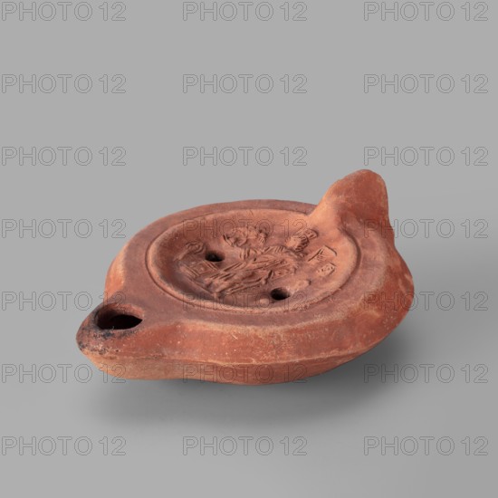 Lamp decorated with a representation of the goddess Roma with an eagle and a palm branch; at the goddess's foot a burning altar and a crab. Roman single-burner oil lamp, 2nd century, Carthage Tunisia. Weight 0.152 kg., A small terracotta oil lamp sits on a light gray surface against a pale gray background, the lamp showing a rounded, flattened body with a raised rim around a shallow central depression that contains a low-relief decorative scene; the lamp has a tapered spout with a darkened rim opening and a tall, rounded handle-like projection at the opposite end. The lamp's surface is primarily reddish-brown with variations of lighter and darker brown tones and small areas of blackened discoloration near the spout, and the texture appears matte and slightly rough with subtle wear and pitting.