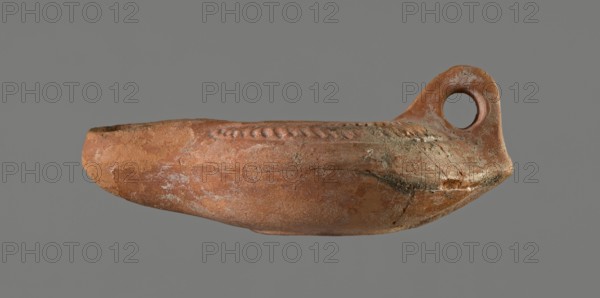 Oil lamp, 5th century, Carthage, oil lamp, height 5.3 cm, width 8.7 cm, depth 13.6 cm, A single worn clay vessel in shades of reddish-brown with areas of lighter tan and grayish-white encrustation is shown against a neutral gray background; the vessel has an elongated, boat-like body with a rounded bottom and a raised rim with a decorative braided or rope-like ridge along the top edge, and a looped handle with a circular opening at one end that shares the same textured, weathered surface and coloration.