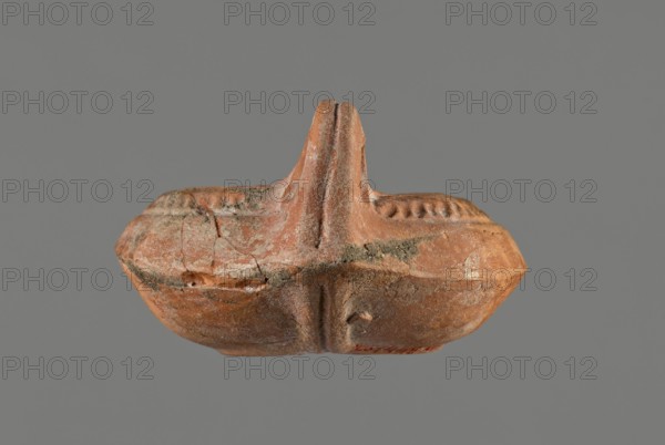 Oil lamp, 5th century, Carthage, oil lamp, height 5.3 cm, width 8.7 cm, depth 13.6 cm, A small, worn terracotta object in shades of reddish-brown and orange sits against a gray background; it has a rounded, boat-like body with a central raised vertical ridge forming a split peak, and a textured rim with a pattern of small indentations. The surface shows areas of lighter beige and pale gray weathering and deposits, with visible cracks and rough patches, and the underside displays smoother, slightly darker brown tones. Overall the piece appears aged with uneven coloration including reddish-brown, orange, beige, pale gray, and darker brown.