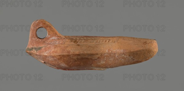 Oil lamp, 5th century, Carthage; oil lamp; height 5.3 cm, width 8.7 cm, depth 13.6 cm, A weathered clay vessel with a rounded base and an integrated looped handle at one end, shown against a plain gray background; the surface shows rough, uneven textures with chips and pitted areas and a few shallow decorative grooves along the rim. The visible colors are terracotta orange, light brown, pale beige, and small patches of off-white and darker brown.