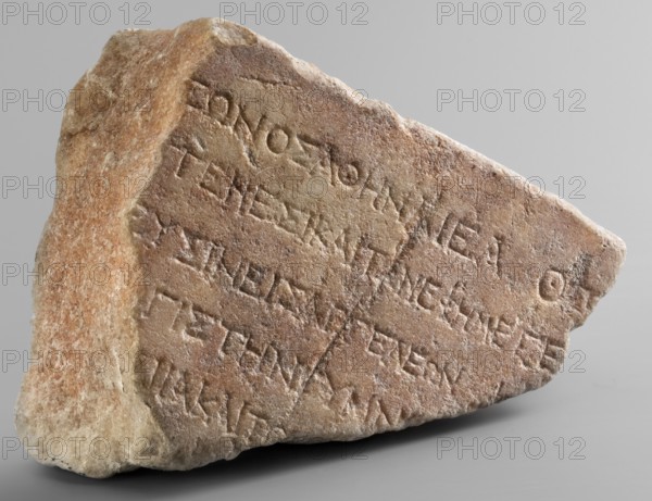 Honorific inscription of Theagenes, son of Theon, Greek inscriptions, marble block covered with a pink patina, fragmentarily preserved. Only the top edge is original. 5 lines of Greek text visible. 115–110 BC, Egypt, Pharbaitos, inscription Greek. Height 15.2 cm, width 21 cm, depth 8 cm, weight 3.70 kg., A roughly triangular fragment of stone with a textured, weathered surface sits against a plain gray background; the stone shows a range of colors including beige, tan, light brown, reddish-brown, and hints of pale cream, with areas of smoother, worn faces and rougher, jagged edges. The front face is flat and slightly angled, displaying shallow, incised markings arranged in several horizontal lines, while the opposite side is more irregular and uneven. The stone’s surface has subtle variations in tone and small pits and scratches, and the base rests on the surface casting a soft shadow. The overall appearance conveys an aged, natural piece of rock with carved linear impressions and a wa...
