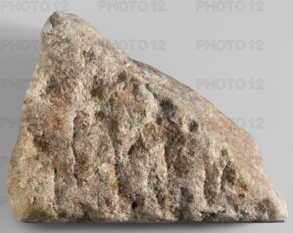 Honorific inscription of Theagenes, son of Theon; Greek inscriptions on a marble block covered with a pink patina, fragmentarily preserved; only the top edge is original. Five lines of Greek text visible. Dated 115–110 BC. Egypt, Pharbaitos locality; place of finding unknown. Inscription Greek. Height 15.2 cm, width 21 cm, depth 8 cm, weight 3.70 kg., A rough, irregularly shaped rock with a triangular profile rests against a plain light gray background; its surface shows a textured, granular appearance with a mix of beige, light brown, tan, cream, white, and small patches of darker gray and muted rust tones, and a few shadowed crevices and pitted areas visible across the face.