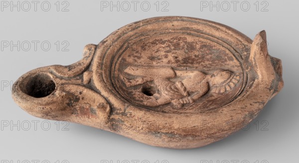 Lamp decorated with a depiction of a gladiator so-called Samnite, unknown Roman workshop, Scen.Gal.Class., gladiators iconography, lighting, oil lamps, single-burner lamp, 2nd half of the 2nd century, Roman Empire 27 BC–476, oil lamp, weight 0.078 kg, An oval, shallow clay object with a rounded spout and raised lip, showing a sculpted reclining figure within a recessed central area; the surface is textured with worn patches and irregularities, and there are decorative raised ridges around the inner rim and a raised element at the opposite end from the spout. The visible colors are shades of brown, terracotta, and darker brown or blackened areas, with some lighter beige highlights.