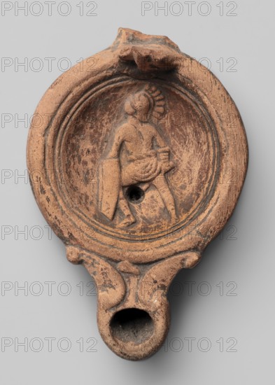 Lamp decorated with a depiction of a gladiator so-called Samnite, unknown Roman workshop, Scen.Gal.Class., gladiators iconography, lighting, oil lamps, single-burner lamps, 2nd half of the 2nd century, Roman Empire 27 BC–476, oil lamp, weight 0.078 kg, An oval terracotta object in shades of reddish-brown and dark brown with weathered surface texture shows a recessed circular field containing a low-relief figure wearing a draped garment and a helmet-like headpiece, standing with one leg forward and holding a rectangular shield and a short object; concentric raised rings surround the recessed field, and there is a central round hole within the scene. The rim and body exhibit worn areas and darker patina, with a small protrusion at the top and a narrow elongated spout at the bottom that forms a rounded opening; the lower handle area features molded scroll-like shapes and similar worn coloration.