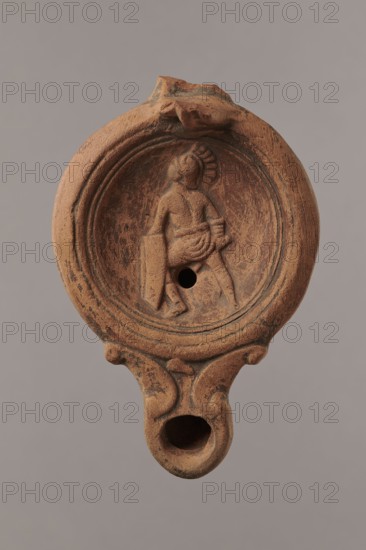 Lamp decorated with a depiction of a gladiator so-called Samnite, unknown Roman workshop, scenographic gladiator class, gladiators iconography, lighting, oil lamp, single-burner lamp, 2nd half of the 2nd century, Roman Empire 27 BC–476, oil lamp, weight 0.078 kg, A clay oil lamp in shades of reddish-brown and tan with weathered surface texture, showing a round central reservoir framed by concentric raised rings and a circular rim; inside the reservoir is a low-relief human figure wearing a helmet-like headpiece and a draped garment, seated and holding a rectangular object, with a round hole set near the figure; the lamp’s nozzle extends downward in a tapered form with a rounded opening and sculpted curved supports connecting it to the body, and the top edge shows an irregular chipped area.