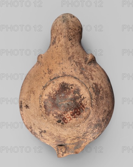 Lamp decorated with a depiction of a gladiator so-called Samnite, unknown Roman workshop, Scen.Gal.Class., gladiators iconography, lighting, oil lamp, single-burner lamp, 2nd half of the 2nd century, Roman Empire 27 BC–476, oil lamp, weight 0.078 kg, A worn, rounded clay object with an elongated top and a small protrusion at the bottom sits against a neutral background; its surface shows a weathered texture with rough patches and flaking, and the predominant colors visible are tan, brown, and darker brown spots with hints of gray and muted reddish areas.
