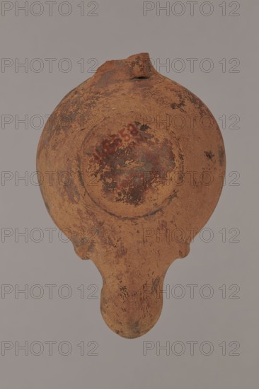 Lamp decorated with a depiction of a gladiator so-called Samnite, unknown Roman workshop, Scen.Gal.Class., gladiators iconography, lighting, oil lamps, single-burner lamp, 2nd half of the 2nd century, Roman Empire 27 BC–476, oil lamp, weight 0.078 kg, A roughly round, flattened ceramic object with a short spout and a teardrop-shaped protrusion, shown from above against a plain gray background; the surface is textured and worn with a predominantly reddish-brown clay color, with areas of darker brown, blackish mottling, and traces of reddish and greenish discoloration, and visible patches of roughness and small chips.